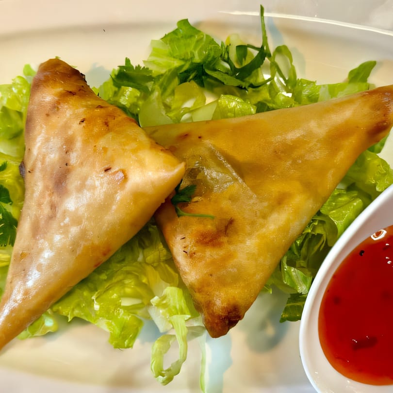 Two golden-brown Bangladeshi samosas on a bed of fresh lettuce, with a side of red dipping sauce.