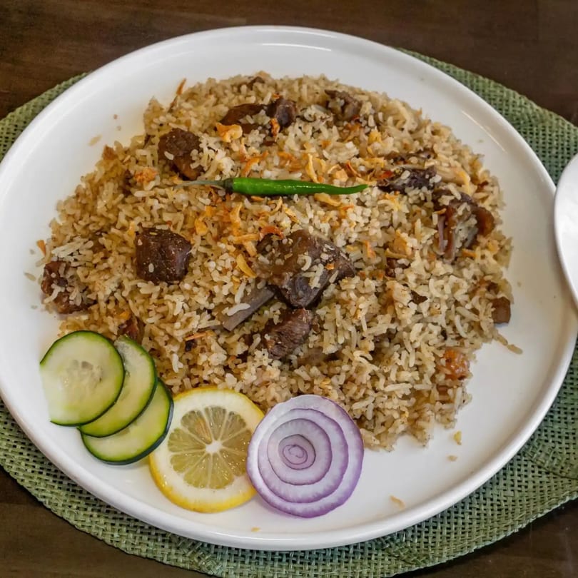 A plate of Bangladeshi beef biryani with a green chili, cucumber, lemon, and red onion.