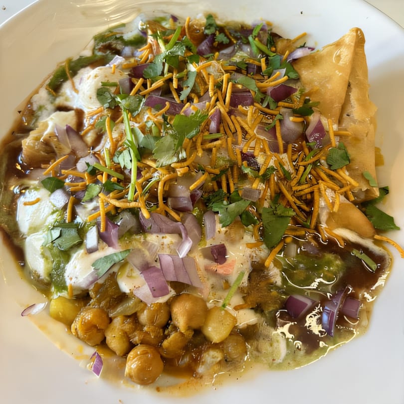 A Bangladeshi samosa chaat with chickpeas, yogurt, sauces, red onions, and crispy noodles.