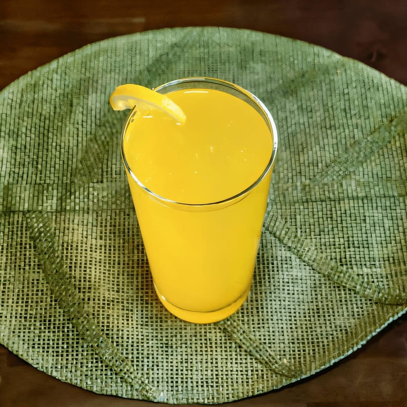 A glass of bright yellow juice with a lemon slice, on a green woven placemat.