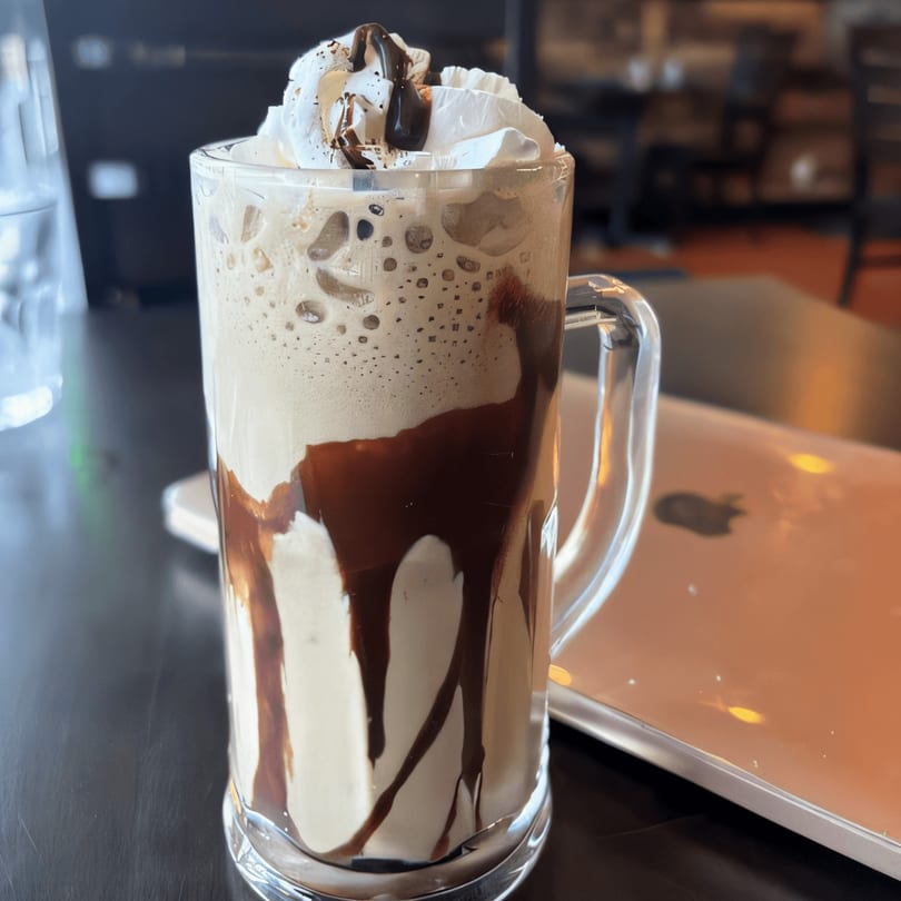 A tall glass of iced coffee with whipped cream and chocolate drizzle on a dark table.